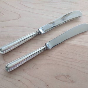 Puede incluir: Dos cuchillos de mantequilla de plata con mangos acanalados. Los cuchillos están dispuestos sobre una superficie de madera clara.