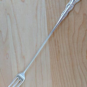 Könnte beinhalten: Eine silberne Cocktailgabel mit einem verzierten Griff und drei Zinken. Die Gabel liegt auf einer hellbraunen Holzoberfläche.