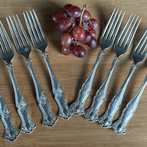 May include: Eight silver forks with ornate designs, arranged in a semi-circle on a wooden surface. The forks have a polished finish and intricate details.