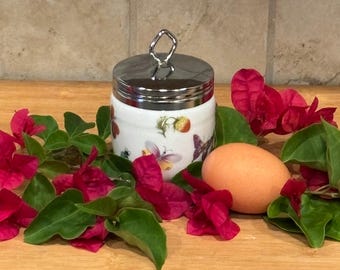 May include: A white and silver egg cup with a butterfly and strawberry design, topped with a silver lid and handle. The egg cup is surrounded by vibrant red flowers and green leaves, with a single egg.