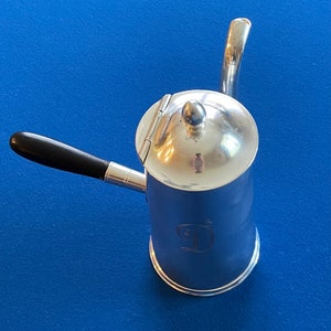 May include: Silver coffee pot with a black handle and a hinged lid. The pot has a decorative design on the side.