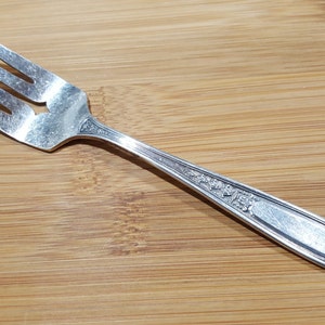 Könnte beinhalten: Eine silberne Gabel mit einem verzierten Griff. Die Gabel liegt auf einer Holzoberfläche.