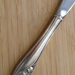 Puede incluir: Cuchillo de mantequilla de plata con un diseño floral en el mango. El mango está decorado con tres flores de lirio de los valles.
