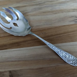 Pode incluir: Uma colher de servir de prata com um design ornamentado e uma cabeça perfurada de três pontas.