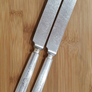 May include: Two silver dinner knives with ornate handles on a wooden surface.