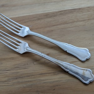 May include: Two silver forks with ornate handles. The forks are lying on a wooden surface.