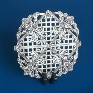 May include: A silver-plated, decorative, openwork, square-shaped serving tray with a grid pattern and ornate scrollwork. The tray is on a black stand.
