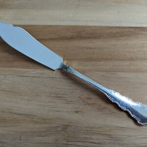 May include: A silver butter knife with an ornate handle and a smooth, slightly curved blade.