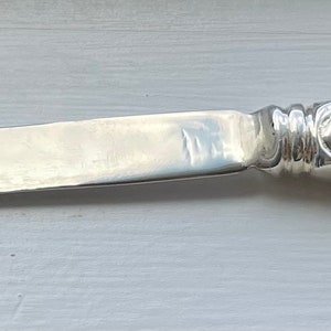May include: A silver letter opener with an ornate handle. The blade is long and slender, and the handle features a detailed floral and scroll design. The letter opener is made of a shiny, reflective metal.