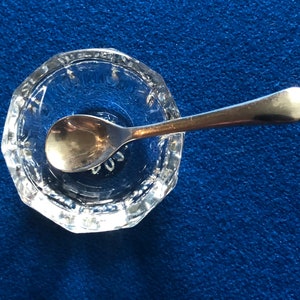 May include: A clear glass salt cellar with a silver spoon inside. The cellar has a decorative, octagonal rim.
