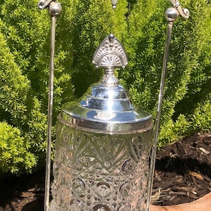 May include: A silver-plated glass sugar dispenser with a cut glass design and a decorative top. The dispenser is on a stand with a silver-plated base and a decorative arch.