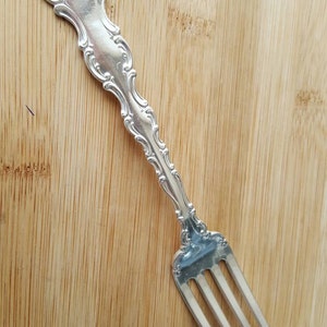 Puede incluir: Un tenedor de plata con un mango adornado y tres dientes. El mango tiene un diseño decorativo y está grabado con iniciales.