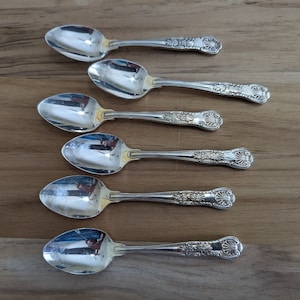May include: Set of six silver teaspoons with ornate handles. The spoons have a polished finish and are arranged in a row on a wooden surface.