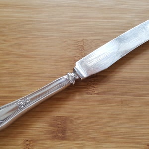 May include: A silver butter knife with an ornate handle. The knife is lying on a wooden surface.