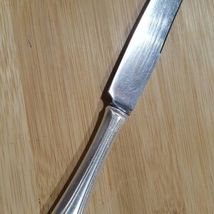 Pode incluir: Uma faca de jantar de prata com uma lâmina ligeiramente curva e um cabo detalhado. O cabo tem um design decorativo e a letra "J" gravada na ponta.
