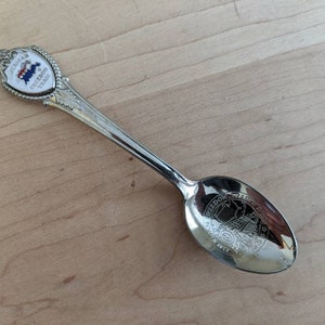 Puede incluir: Una cuchara plateada con mango y cuenco decorativos. El mango presenta un emblema en forma de escudo con las palabras "AMERICAN FREEDOM TRAIL" y un diseño rojo, blanco y azul. El cuenco tiene un diseño grabado.