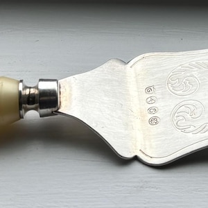 May include: A silver cake server with an ornate design and a cream-coloured plastic handle.