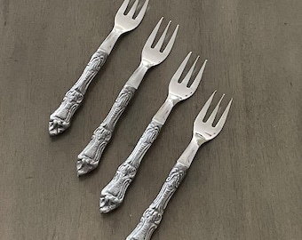 May include: Set of four silver-toned ornate forks with decorative handles. The forks are arranged in a row on a brown wooden surface.