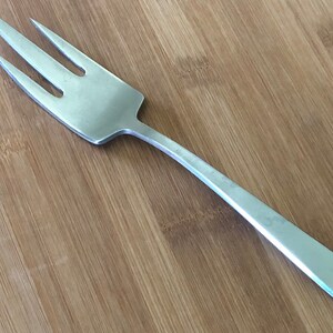 以下が含まれることがあります： 長い細い柄のついた銀のフォーク。フォークは4本の歯があり、木製の表面の上に置かれています。