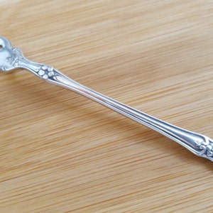 Könnte beinhalten: Eine silberne Gabel mit drei Zinken, die ein dekoratives Blumenmuster am Griff und in der Nähe der Zinken aufweist. Die Gabel liegt auf einer hellbraunen Holzoberfläche und zeigt ihr elegantes Design.