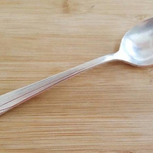Puede incluir: Una cucharadita de plata con un mango decorativo. La cuchara está sobre una superficie de madera.