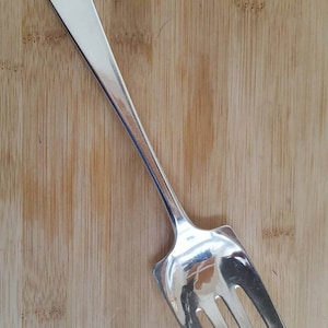 May include: A silver serving fork with a long, slender handle and three tines. The fork is on a wooden surface.
