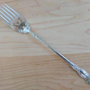 Könnte beinhalten: Eine silberne Serviergabel mit einem verzierten Griff und einem dekorativen durchbrochenen Design. Die Gabel liegt auf einer hellholzfarbenen Oberfläche.