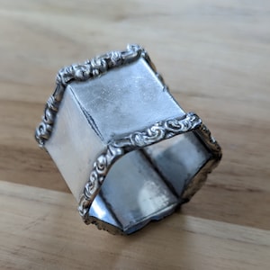 Puede incluir: Anillo de servilleta de plata con un diseño hexagonal adornado. El anillo tiene un acabado liso y pulido y detalles intrincados alrededor de los bordes.