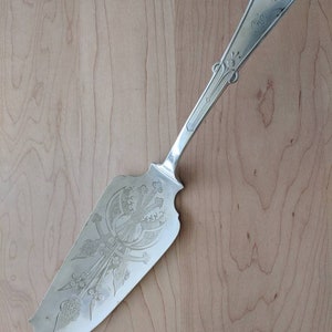 Puede incluir: Un servidor de pastel de plata con un diseño ornamentado. El mango está decorado con un motivo floral y la hoja tiene un patrón detallado.
