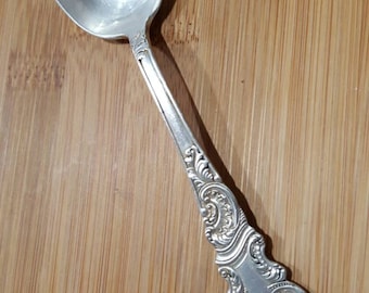 May include: A silver teaspoon with an ornate, detailed handle. The spoon has a smooth, polished finish.