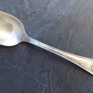 May include: A silver teaspoon with an ornate design on the handle. The handle features a raised image of a person's profile.