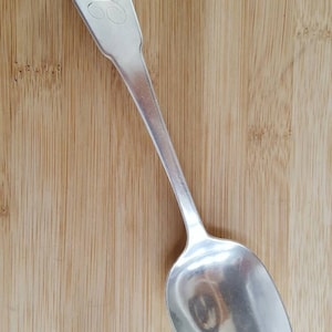 May include: A silver spoon with a shell-shaped handle and an engraved design. The spoon is on a wooden surface.