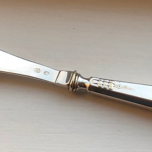 Puede incluir: Un cuchillo de mantequilla de plata con un mango decorativo. El mango tiene un patrón de pequeños círculos y un círculo más grande en el extremo.