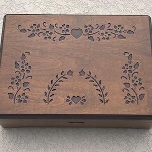 Puede incluir: Una caja de madera con un acabado marrón oscuro y tallas florales intrincadas. La caja tiene una tapa con bisagras y un diseño de corazón decorativo.