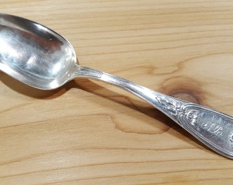 May include: A silver spoon with an ornate handle and the inscription "ELB" on the back. The spoon has a rounded bowl and a polished finish.