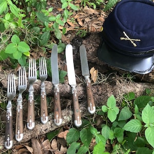 May include: A set of six antique silver forks and three antique silver knives with wooden handles. The forks and knives are arranged on a wooden surface with green foliage in the background. A blue military cap with a crossed-gun insignia is also visible in the image.