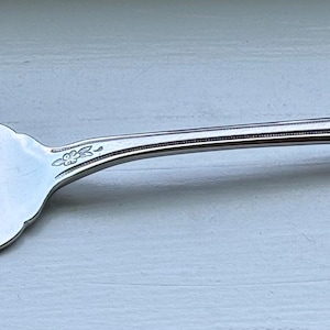 Op de afbeelding: Een zilverkleurige metalen serveervork met drie tanden en een centrale ruitvormige uitsparing in de kop. De steel is slank met een uitlopend uiteinde en is voorzien van verfijnde gegraveerde florale krulmotieven op de hals en steel.