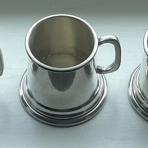 Könnte beinhalten: Fünf silberne Zinnbecher mit abgerundetem Boden und Henkel. Die Becher sind in einer Reihe auf einer weißen Oberfläche angeordnet.