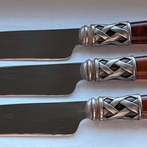 May include: Three butter knives with stainless steel blades and brown, marbled handles. The handles are connected to the blades with a silver, woven design. The knives are arranged horizontally on a white surface.
