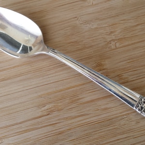 Puede incluir: Una cuchara de plata con un diseño floral ornamentado en el mango. La cuchara está sobre una superficie de madera.
