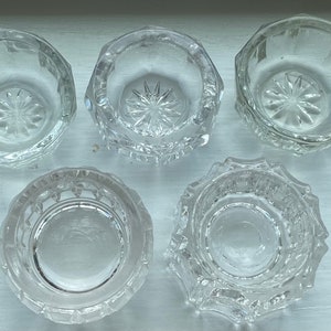 May include: Five clear glass salt cellars with a decorative pattern on the bottom. The salt cellars are all the same size and shape.