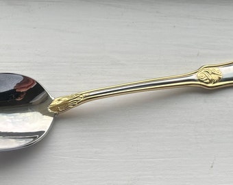 May include: A silver spoon with a gold-plated handle featuring an ornate design. The spoon has a rounded bowl and a long, slender handle.