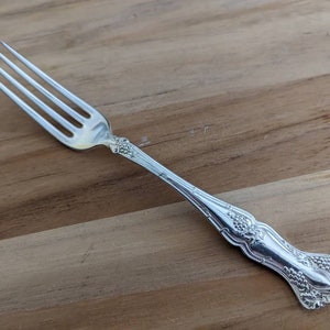 May include: A silver fork with an ornate handle featuring a grapevine design. The fork has four tines and a polished finish.