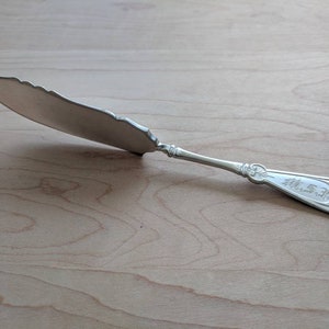 Puede incluir: Un cuchillo de mantequilla de plata con un mango adornado y un diseño decorativo en la hoja. El mango tiene la inscripción "H.S."