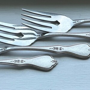 Op de afbeelding: Een set van vier zilverkleurige metalen vorken, diagonaal gerangschikt. Elke vork heeft een kop met vier tanden en een handvat dat bij de hals is versierd met een klein, gegraveerd bloemmotief en eindigt in een afgeronde, puntige vorm.