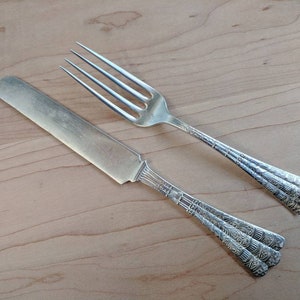 Puede incluir: Un tenedor y un cuchillo de plata con diseños ornamentados en los mangos. El tenedor tiene cuatro dientes y el cuchillo tiene una hoja puntiaguda.
