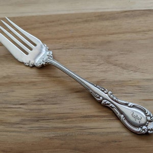 Puede incluir: Un tenedor de plata con un mango adornado y un monograma "J" en la parte posterior. El tenedor está sobre una superficie de madera.