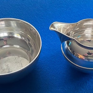 Könnte beinhalten: Ein silbernes Sahnegießer- und Zuckergefäß-Set. Der Sahnegießer hat einen runden Körper und einen Griff mit einem dekorativen Knauf. Die Zuckerdose ist rund und hat eine glatte, reflektierende Oberfläche.