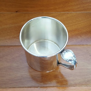 Könnte beinhalten: Eine silberfarbene Tasse mit dekorativem Henkel. Die Tasse ist zylindrisch mit einer weiten Öffnung und einer glänzenden, reflektierenden Oberfläche. Der Henkel hat ein verziertes, florales Design. Die Tasse steht vor einem Holzgrund.