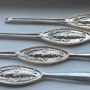 以下が含まれることがあります： 装飾的なディテールが施された、ロブスターの形をした銀製のバターナイフが4本。ナイフは一列に並んでおり、ハンドルは右を向いています。